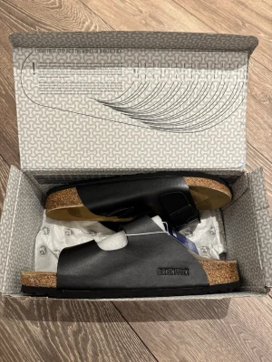 Svarta Birkenstock sandaler - Säljer ett par klassiska svarta Birkenstock sandaler med två justerbara remmar och spännen. Fotbädden är i kork och mocka för extra komfort. Perfekta för dig som vill ha både stil och bekvämlighet. 
