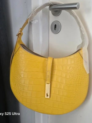 Gul croco handväska från Lauren Ralph Lauren - Cool gul handväska från Lauren Ralph Lauren i croco-mönstrat läder. Väskan har en rundad form, bred axelrem och snyggt guldfärgat spänne med logotyp. Perfekt för dig som vill sticka ut med färg och stil.