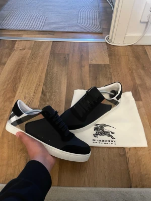 Burberry svart  sneakers - Snygga svarta sneakers från Burberry med klassiskt rutmönster på hälen och vita sulor. Skorna har en stilren design med svarta skosnören och detaljer i mocka och textil. Perfekta för dig som vill ha en exklusiv och trendig look.