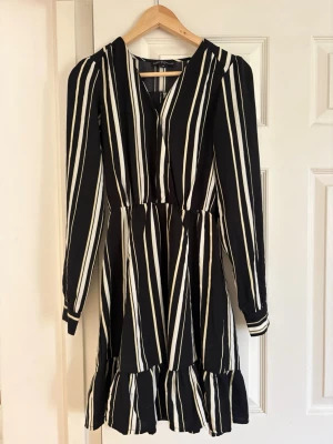 Svartvit randig klänning med volang - Svart klänning med vita vertikala ränder och v-ringning. Långärmad modell med volangkant nedtill och markerad midja. Perfekt för dig som gillar en stilren look med lite edge. Tillverkad i ett mjukt, lätt material som känns skönt mot huden.