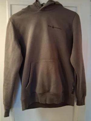 Hoodie från Sail Racing - En  hoodie från Sail Racing med klassisk huva och stor magficka. Diskret logga på bröstet samt uppe på luvan. Färgen är grå/grön med lite solblekt framtill på luvkanten.