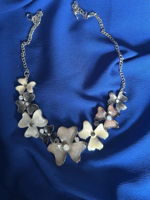 Blommigt halsband med pärldetaljer - Snyggt halsband med stora blommor i beige, vitt och brunt, dekorerade med pärlor och små stenar. Kedjan är silverfärgad och blommorna har en glansig finish. Perfekt för att ge din outfit en unik touch.