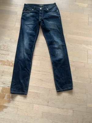 Mörkblå slim fit jeans från Philipp Plein - Säljer ett par mörkblå slim fit jeans från Philipp Plein med snygga slitningar och silverfärgade nitar vid fickorna. Klassisk femficksmodell med kontrastsömmar och knappstängning. Perfekt för dig som gillar en cool och trendig look.