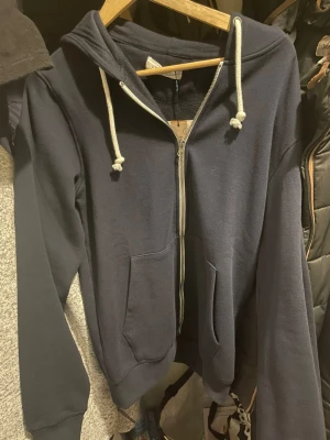 Mörkblå zip-hoodie med huva - En helt ny mörkblå hoodie med dragkedja framtill, två stora fickor och vit dragsko i huvan. Tröjan har ribbade muddar vid ärmslut och nederkant. Perfekt för chill dagar och enkel att matcha med jeans eller joggers.