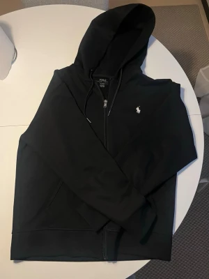 Svart hoodie från Polo Ralph Lauren - Säljer en svart hoodie från Polo Ralph Lauren med dragkedja och huva. Klassisk design med vit logotyp på bröstet och praktiska fickor framtill och Materialet är mjukt och bekvämt. 