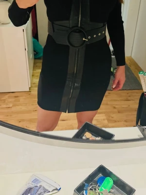 Svart klänning från Guess Marciano - Svart långärmad klänning från Guess Marciano med markerad v-ringning, skinndetaljer och dekorativ dragkedja framtill. Klänningen har ett brett bälte med stor rund spänne som accentuerar midjan. Materialet är stickat och ger en slim passform.