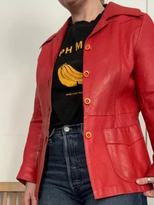 Röd skinnjacka med knappar, 36 - Cool röd skinnjacka med klassisk krage och fyra snygga knappar framtill från Fritala. Jackan har två stora fickor och raka ärmar. Jackan är väl använd, men slitaget bidrar till dess charm.