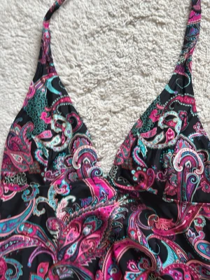 Svart tröja, badtyg. med paisleymönster - Snygg tröja med färgstarkt paisleymönster i rosa, turkos och vitt på svart bakgrund. Halterneck-modell med knytband i nacken och v-ringad front. Perfekt för strandhäng och poolparty.