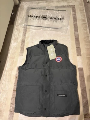 Mörkgrå dunväst från Canada Goose - Snygg mörkgrå dunväst från Canada Goose med klassisk logotyp på bröstet och två stora fickor framtill. Västen har hög krage och knäppning med svarta knappar. Perfekt för lager-på-lager och håller dig varm under kyliga dagar. Pris kan diskuteras