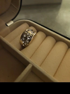 Guldfärgad ring med gnistrande stenar - Säljer en bred guldfärgad ring med flera gnistrande klara stenar i olika storlekar. Ringen har en rundad form och är riktigt eye-catching, perfekt för dig som gillar att sticka ut. Levereras i ett smyckesetui.