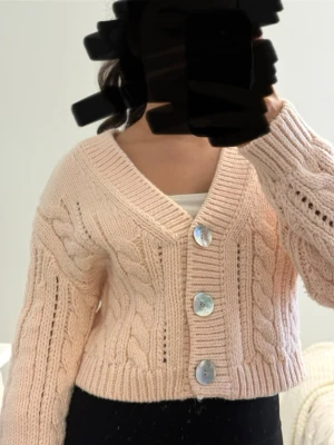 Ljusrosa kabelstickad kofta - Superfin ljusrosa kabelstickad kofta med stora pärlemorknappar och djup v-ringning. Koftan är kortare i modellen och har breda ribbade muddar. Perfekt att slänga över en topp för en chill look. Materialet är tjockt och mjukt, troligtvis akryl eller ullblandning.