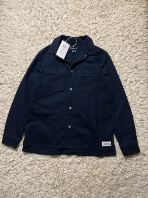 Mörkblå skjorta från Name It - Snygg mörkblå skjorta från Name It med klassisk krage och två bröstfickor. Skjortan har knappar framtill och är gjord i ett mjukt bomullsmaterial. Perfekt att slänga på över en t-shirt för en avslappnad look.