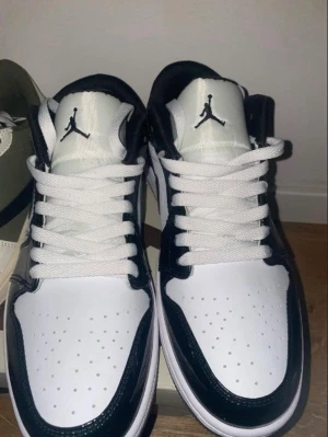 Air Jordan 1 Low svart/vit - Säljer ett par Air Jordan 1 Low i svart och vitt. Skorna har klassisk låg siluett, vit tåbox med perforeringar, svarta detaljer i blankt läder och vita skosnören. Ikoniska Jumpman-loggan på plösen och Swoosh på sidan. Perfekta för dig som gillar streetwear och sneakerskultur.
