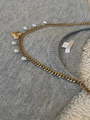 Guldhalsband med pärlor och hjärta - Snyggt halsband i guldfärgad rostfritt stål med små vita pärlor och ett större hjärthänge. Kedjan har en chunky look och pärlorna hänger jämnt längs hela halsbandet. Perfekt accessoar för att lyfta vilken outfit som helst.