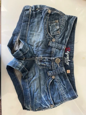 Snygga Jeansshorts  - Säljer ett par blå denimshorts från Reporter med klassisk femficksdesign, dubbla knappar och snygga detaljer på fickorna. Shortsens material är bomull och de har en lågmidjad passform. Perfekta för sommaren! 