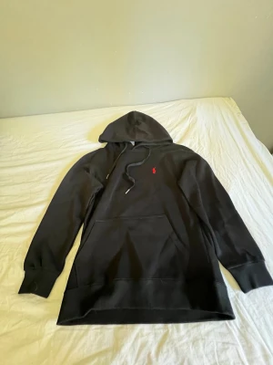 Svart hoodie från Polo Ralph Lauren - Svart hoodie med huva och dragsnören från Polo Ralph Lauren. Klassisk röd broderad logga på bröstet och stor magficka framtill. Mjuk bomullskänsla och ribbade muddar vid ärmslut och nederkant. Perfekt för en chill look.
