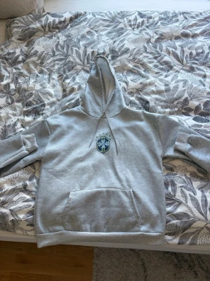 Grått mjukisset med tryck - Säljer ett grått mjukisset med hoodie och byxor. Hoodien har magficka, dragsko i huvan och ett tryckt emblem på bröstet. Byxorna har resår i midjan och samma emblem på låret. Setet är gjort i mjukt sweatshirtmaterial, perfekt för chill dagar. Pris kan diskuteras!