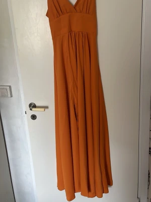Lång orange klänning med v-ringning - Snygg långklänning i en stark orange färg med v-ringad hals och axelband. Klänningen har ett luftigt fall och markerad midja, perfekt för sommarens alla tillfällen. Tillverkad i ett mjukt och lätt material som ger en skön känsla.