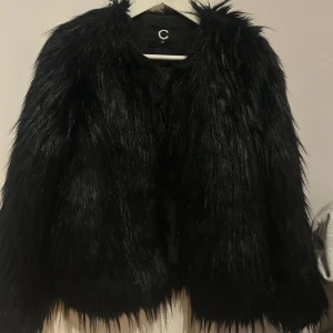 Svart pälsjacka  - Cool svart pälsjacka från Cubus med lång, fluffig syntetpäls och rund halsringning. Jackan har en rak passform och är perfekt för att skapa en edgy look under vintern. Snygg att styla med jeans eller klänning.