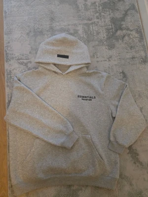 Grå Essentials hoodie från Fear of God - Säljer en grå hoodie från Fear of God Essentials med svart logotyp på bröstet. Hoodien har en stor huva, ribbade muddar och en klassisk känguruficka framtill. Perfekt för dig som gillar streetwear och enkel stil.
