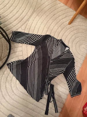 Svartvit omlottblus med knyt - Säljer en svartvit omlottblus med knyt i sidan och v-ringad hals. Blusen har ett randigt mönster och långa ärmar. Perfekt för dig som gillar stilrena plagg med lite edge. Materialet känns mjukt och lätt, troligen polyester eller bomull.