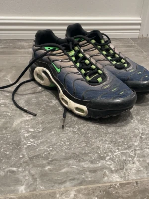 Nike Air Max Plus blå och grön - Nike Air Max Plus sneakers med blå mesh och grå detaljer, gröna accenter och svart sula. TN-logga på undersidan och klassisk vågig design. Skorna har svarta snören och gröna inslag på sulan. Perfekt för dig som gillar streetstyle och ikoniska modeller.