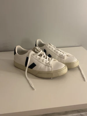 Vita Veja sneakers med svarta detaljer - Snygga vita sneakers från Veja med svarta V-loggor på sidorna och svarta detaljer vid hälen. Skorna har klassisk låg profil, vita snören och en beige sula. Tillverkade i läder med en stilren och tidlös design. Köpt på Vinted men passade ej i storleken. Allmänt väldigt bra skick, dock lite defekter på sulan som syns på bilderna.