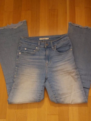 Ljusblå Levi's bootcut jeans - Säljer ett par ljusblå Levi's jeans med flare form och slitna, råa kanter längst ner på benen. Jeansen har klassisk femficksdesign, knappgylf och tydliga slitningar framtill för en vintagekänsla. Tillverkade i denim med Levi's ikoniska läderpatch bak.