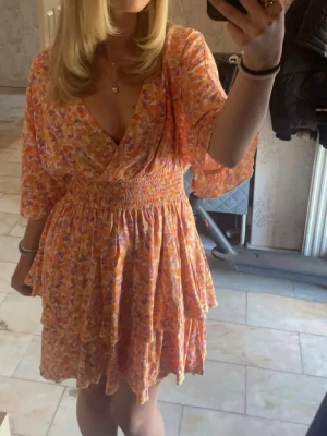 Blommig omlottklänning i orange och lila - Säljer en söt omlottklänning med blommigt mönster i orange, lila och rosa toner. Klänningen har v-ringning, vida trekvartsärmar och smock i midjan för en snygg passform. Kjolen är volangig och luftig, perfekt för varma dagar.