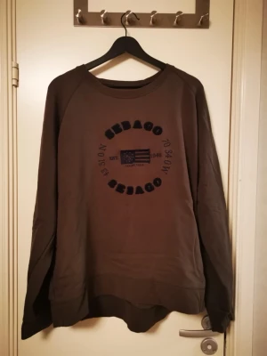 Brun sweatshirt från Sebago - Mysig brun sweatshirt från Sebago med tryckt logga och amerikansk flagga på bröstet. Tröjan har rund halsringning och långa ärmar, perfekt för en avslappnad stil. Tillverkad i mjukt material som känns skönt mot huden.