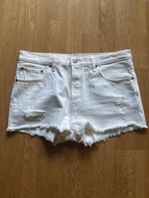 Vita denimshorts med fransig kant - Säljer ett par vita denimshorts med rå, fransig kant och slitna detaljer framtill. Klassisk femficksmodell med knapp och dragkedja. Perfekta för sommaren och ger en avslappnad vibe.