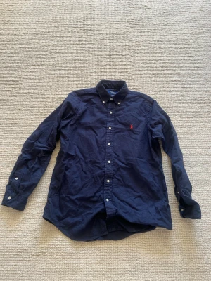 Mörkblå skjorta från Polo Ralph Lauren - Klassisk mörkblå skjorta från Polo Ralph Lauren med button-down krage och röd broderad logga på bröstet. Skjortan har lång ärm, vita knappar och är tillverkad i bomull. Perfekt för en clean och stilren look.
