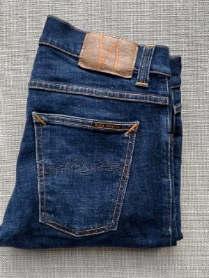 Blå jeans från Nudie Jeans - Säljer ett par klassiska blå jeans från Nudie Jeans med snygga kontrastsömmar och läderpatch bak. Jeansen har fem fickor och en slim fit-modell. Materialet är kraftig denim och detaljerna är typiska för märket. Perfekt för dig som gillar stilrena och tidlösa jeans. Priset är inte hugget i sten. Bara att höra av sig vid minsta fråga