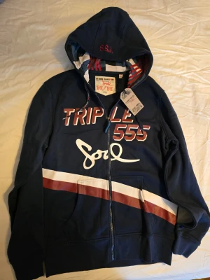 Blå hoodie från Triple 555 Soul - Snygg mörkblå hoodie med dragkedja från Triple 555 Soul. Den har huva med röd- och vitrandigt foder, tryck på bröstet och diagonala rödvita ränder över magen. Perfekt för dig som gillar streetwear och coola detaljer.