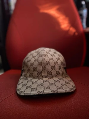 Beige keps från Gucci med GG-mönster - Säljer en beige keps från Gucci med det klassiska GG-monogrammönstret över hela kepsen. Kepsen har böjd skärm och en detalj med gröna och röda ränder på sidan. Tillverkad i canvasmaterial som ger en exklusiv känsla.