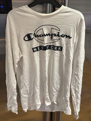 Vit långärmad Champion t-shirt - Vit långärmad t-shirt från Champion med stor svart logga och texten 'New York' på bröstet. Tillverkad i mjuk bomull med avslappnad passform och rund halsringning. Perfekt för en casual streetwear-look.