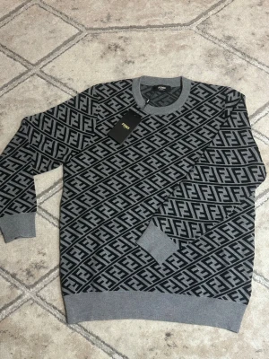 Grå Fendi tröja med FF-mönster - Fendi FF Motif Stickad Tröja - Grå/Svart Stickad tröja från Fendi i bomull, lämplig som ett värmande lager under sena sommarkvällar. Plagget har ett genomgående FF-mönster i jacquardstickning.🌘💫
