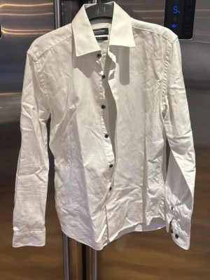 Vit slim fit skjorta från Dressmann - Vit skjorta från Dressmann i premium bomull med slim fit-passform. Klassisk krage, svarta knappar och långa ärmar med knapp vid manschetten. Perfekt för dig som gillar en stilren och modern look.