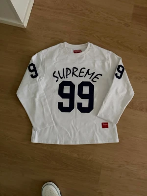 Vit långärmad topp från Supreme - Supreme långärmad topp i vit bomull med stora svarta siffror '99' och Supreme-tryck på bröstet. Siffran 99 finns även på båda ärmarna. Klassisk crewneck och rakt snitt, perfekt för dig som gillar streetwear.