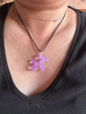 Lila halsband med ballonghund - Säljer ett halsband med ett unikt lila hänge i form av en ballonghund. Hänget är gjort av plast och sitter på ett svart snöre. Perfekt för dig som gillar lekfulla och färgglada accessoarer.