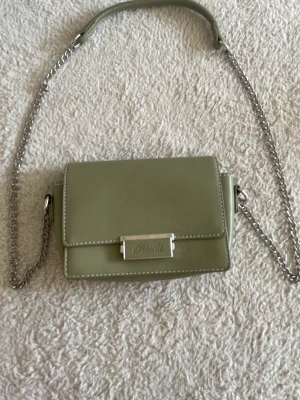 Grön axelväska från Puccini - Stilren axelväska i ljusgrönt från Puccini med silverfärgad kedja och metallspänne. Väskan har en rektangulär form och är tillverkad i slätt syntetmaterial med vita sömmar. Perfekt för dig som gillar minimalistisk design och trendiga accessoarer.