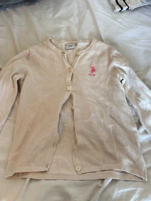 Beige kofta från U.S. Polo Assn. - Säljer en beige kofta från U.S. Polo Assn. med lång ärm och rund hals. Koftan har knappar framtill och ett rosa broderat logotyp på bröstet. Materialet är stickat och mjukt, perfekt för lager-på-lager och chill stil.
