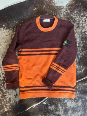 Stone Island stickad tröja röd/orange - Cool stickad tröja från Stone Island i vinröd och orange med kontrasterande ränder och rund hals. Tröjan har lång ärm och klassisk Stone Island patch på vänster ärm. Perfekt för dig som gillar streetwear och snygga detaljer.