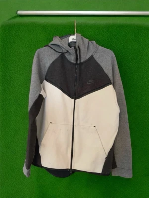 Nike Tech Fleece hoodie grå/vit - Nike Tech Fleece hoodie med dragkedja i grått och vitt. Snygg färgblockad design med mörkgrått upptill och ljusgrå ärmar. Klassisk huva och två fickor framtill. Perfekt för en chill och sportig look. Nike-logga på bröstet.