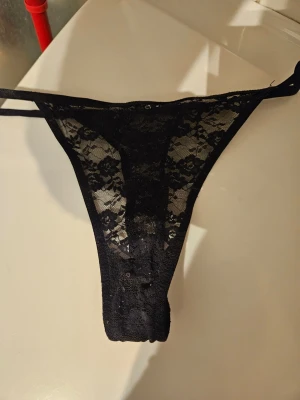 Svarta spets-trosor med strappy detaljer - Säljer ett par svarta trosor i transparent spets med blommönster och tunna strappy band på sidorna. Modellen är en string och materialet är mjuk spets som ger en snygg och bekväm look.