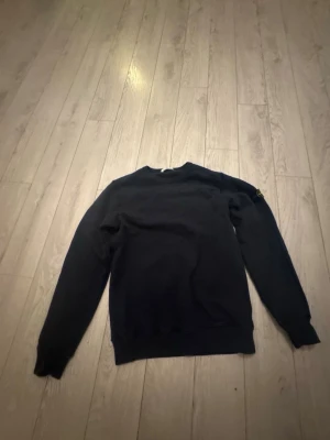 Svart sweatshirt från Stone Island - Säljer en svart sweatshirt från Stone Island med den klassiska loggan på vänster ärm. Tröjan har rund halsringning, ribbade muddar och är tillverkad i mjukt bomullsmaterial. Perfekt för dig som gillar streetwear och stilrena plagg. Storleken är ungefär 170 cm så ungefär xs/s,kom i dm för frågor. Knappt andvänd