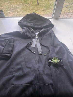 Svart hoodie från Stone Island - Säljer en svart hoodie från Stone Island med dragkedja och huva. Hoodien har ribbade muddar, snörning i huvan och den klassiska Stone Island-loggan på ärmen. Materialet är mjukt och bekvämt, perfekt för en avslappnad look.