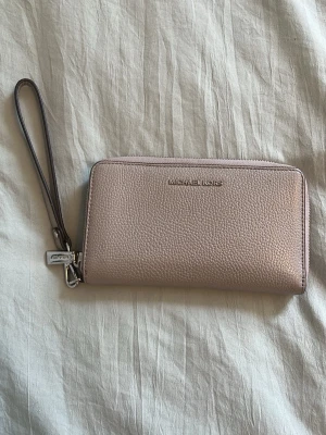 Beige/grå  plånbok från Michael Kors - Säljer en snygg beige/grå plånbok från Michael Kors med struktur i skinn och silverfärgade detaljer. Plånboken har dragkedja runtom och ett praktiskt handledsband. Perfekt för att hålla koll på kort och cash.