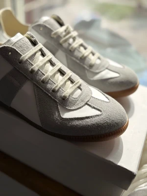 manson margiela - Snygga sneakers i grått och vitt med klassisk design. Ovandelen är i mocka och textil, med vita skosnören och bruna gummisulor. Perfekt för dig som gillar stilrena och tidlösa skor. Skorna har låg profil och passar till många olika outfits.