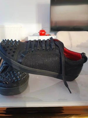 Svarta sneakers med nitar och röd sula - Snygga svarta sneakers från Christian Louboutin med glittrande textil, svarta läderdetaljer och svarta nitar på tån. Insidan är röd och yttersulan har den klassiska röda färgen med mönster. Skorna har svarta skosnören och en låg profil.
